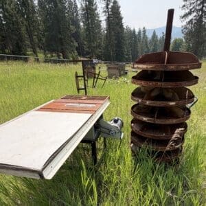 IMG_8934 - Montana Auctions by Kevin Hill Rusty stack of circular metal shelves mounted on a vertical rod beside a metal worktable in a grassy field with trees in the background.
