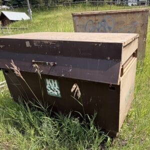 IMG_8937 - Montana Auctions by Kevin Hill A rusty brown dumpster in a grassy field with a small wooden doghouse and graffiti-covered metal cabinet in the background.