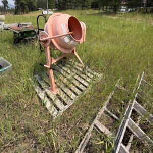 IMG_8948 - Montana Auctions by Kevin Hill Pink portable concrete mixer on a metal frame sits in an overgrown grassy lot with wooden pallets nearby on a sunny day.