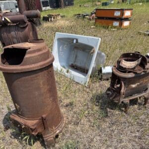 Outdoor scrap yard with rusted metal stoves and a white tub among tall grass.]