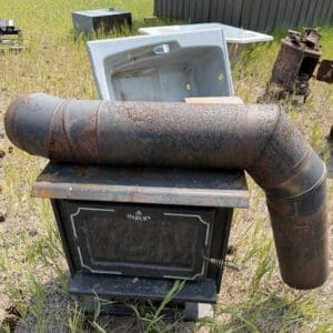 IMG_8954 - Montana Auctions by Kevin Hill Rusty Osburn wood stove outdoors with a curved rusted vent pipe in a grassy, scrap-yard setting.