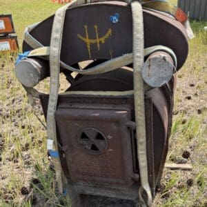 IMG_8955 - Montana Auctions by Kevin Hill Rusty, old industrial furnace-like device strapped with frayed straps, sitting in a grassy field with cones in the background.