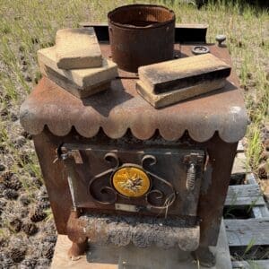 IMG_8956 - Montana Auctions by Kevin Hill Rusty outdoor stove with a decorative yellow dial, bricks on top, set in a dry grassy field outdoors.