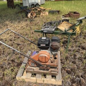 IMG_8967 - Montana Auctions by Kevin Hill Rusty orange gas-powered engine on a pallet in a grassy salvage yard with trailers and discarded gear in the background.