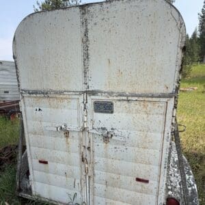 IMG_8979 - Montana Auctions by Kevin Hill Rear view of a rusty white utility trailer with tall curved top, double doors, and weathered metal latches in an overgrown grassy field.
